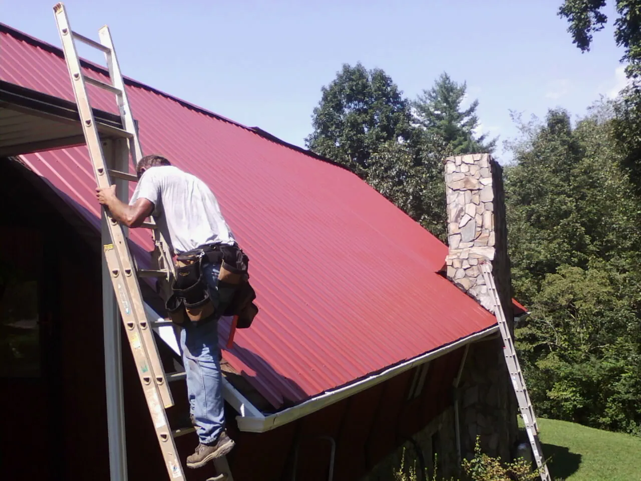 Roofer on a ladder beside a red metal roof and stone chimney — Soffit & Fascia Repair by Roofing Brothers in Cherryville
